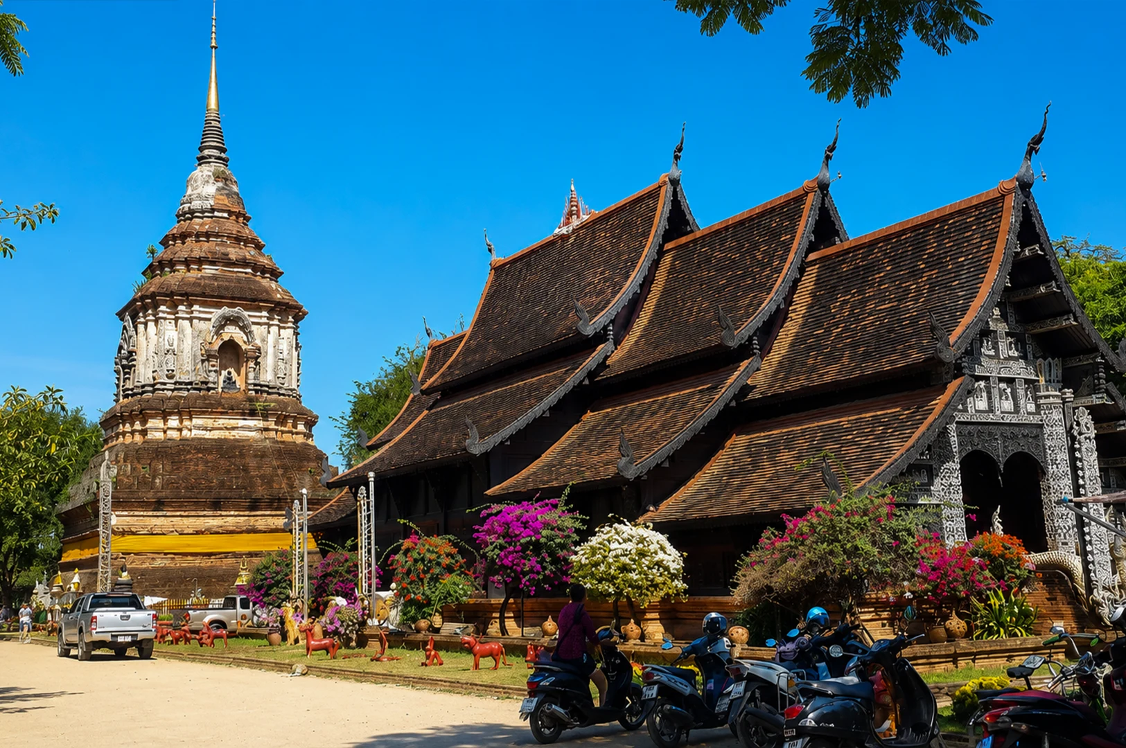 A wide shot of a traditional Northern Thai Buddhist temple complex under a clear, vibrant blue sky. To the left stands an ancient, weathered brick chedi (stupa) with a golden spire, while the main temple building features a multi-tiered, dark wooden roof with ornate gables and intricate silver-colored carvings on its facade. The foreground is bustling with parked motorbikes and colorful bougainvillea bushes in pink and white, while a few silver pickup trucks are parked on the dirt path to the side.