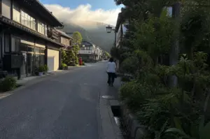 A person wearing a white shirt and dark pants walks away from the camera down a quiet, paved street in a traditional Japanese town during the golden hour. To the left, dark wooden two-story buildings with large glass windows and closed shutters line the road, while the right side features lush green trees and a stone-lined drainage ditch running parallel to the sidewalk. In the background, mist and soft morning sunlight roll over a forested hill, casting a warm, hazy glow over the narrow street and the distant, white-walled buildings.