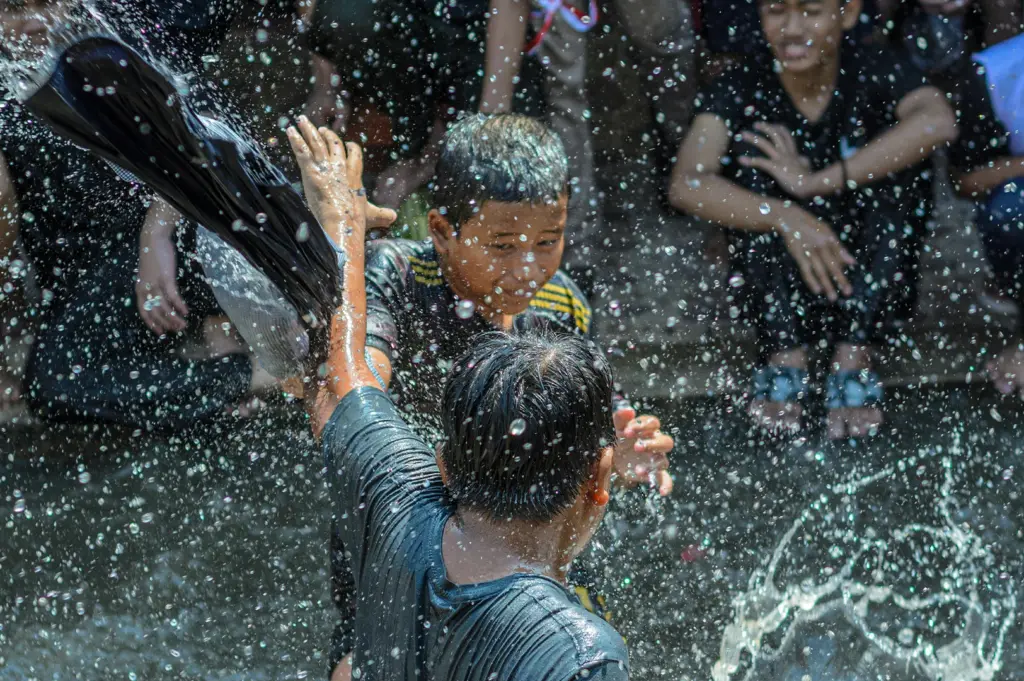 A dynamic, close-up shot of two young boys engaged in a joyful water fight. The boy in the foreground is seen from behind, his black shirt soaked through as he raises a hand to shield himself. Facing him, another boy with a focused expression prepares to splash or dodge. Countless crystalline water droplets are frozen in mid-air, creating a glittering, chaotic texture that fills the frame. In the blurred background, other children sit and watch the play.