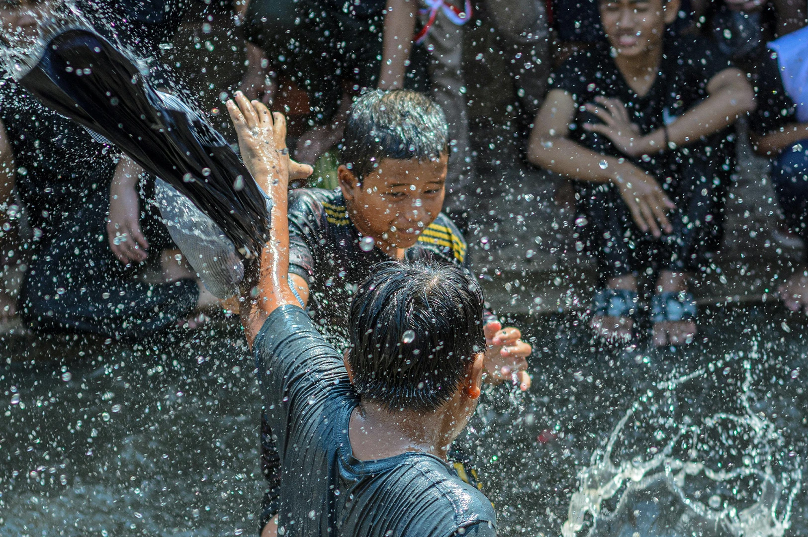 A dynamic, close-up shot of two young boys engaged in a joyful water fight. The boy in the foreground is seen from behind, his black shirt soaked through as he raises a hand to shield himself. Facing him, another boy with a focused expression prepares to splash or dodge. Countless crystalline water droplets are frozen in mid-air, creating a glittering, chaotic texture that fills the frame. In the blurred background, other children sit and watch the play.
