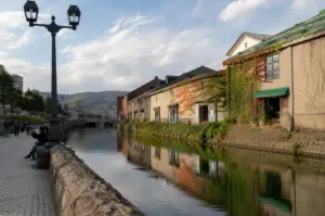 A tranquil view of the Otaru Canal in Hokkaido, Japan, stretches into the distance under a bright, cloud-dappled sky. To the right, a row of historic stone warehouses lined with creeping ivy—some turning a vibrant autumn red—is perfectly reflected in the calm, dark water of the canal. On the left, a cobblestone pedestrian path is bordered by a low stone wall where a few people sit and walk, framed by a vintage-style black lamp post in the foreground. Lush green mountains rise softly in the far background, completing a peaceful scene that blends industrial heritage with natural beauty.