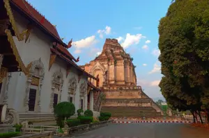 This eye-level outdoor shot captures the historic Wat Chedi Luang in Chiang Mai under a soft late-afternoon sky. To the left stands a white ornate temple building with intricate gold carvings and a tiered red-tiled roof. Dominating the center-right is the massive, weathered stone pagoda (chedi), showing its ancient brickwork and partially crumbled upper structure. Manicured green shrubs line a paved path in the foreground, while a dense, leafy green tree frames the right side of the frame against a backdrop of light blue sky scattered with wispy clouds.