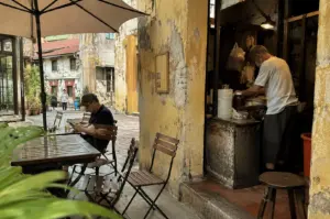 A candid, everyday scene in a narrow, weathered urban alleyway, split between an outdoor seating area and a small, rustic food stall. On the left, a man wearing a dark polo shirt and a baseball cap sits at a wooden slatted table under the shade of a large beige patio umbrella, looking down at his phone. To the right, separated by a thick, distressed yellow pillar with peeling paint, an older man in a white t-shirt and face mask works inside a cramped, dimly lit kitchen, surrounded by stacks of containers and cooking supplies. The background reveals a glimpse of aged multi-story buildings with red-tiled roofs and shuttered windows, while the foreground is framed by soft, out-of-focus green leaves, enhancing the feeling of a quiet, observed moment in a bustling city.