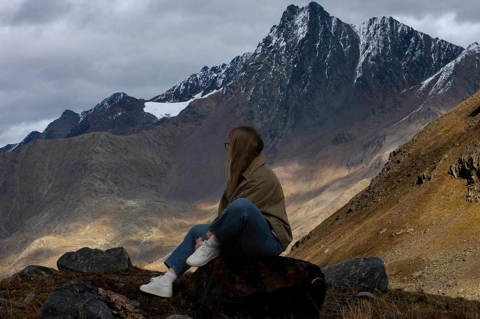 A lone traveler sits on a dark, rugged rock in the foreground of the image titled "You Travel Differently When You Are Not Trying to Impress Anyone.jpg," gazing out at a massive, snow-capped mountain peak under a moody, overcast sky. The individual, seen from behind and dressed casually in a tan jacket, blue jeans, and white sneakers, appears small and contemplative against the vast, rocky landscape that transitions from golden-brown slopes to jagged, dark ridges. Patches of white snow and ice cling to the upper reaches of the mountain, while soft shadows and muted light emphasize the quiet, unposed nature of the moment.