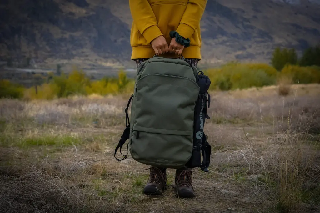 Person in a mustard hoodie and hiking boots holding a green backpack while standing in a grassy mountain landscape during an outdoor travel adventure
