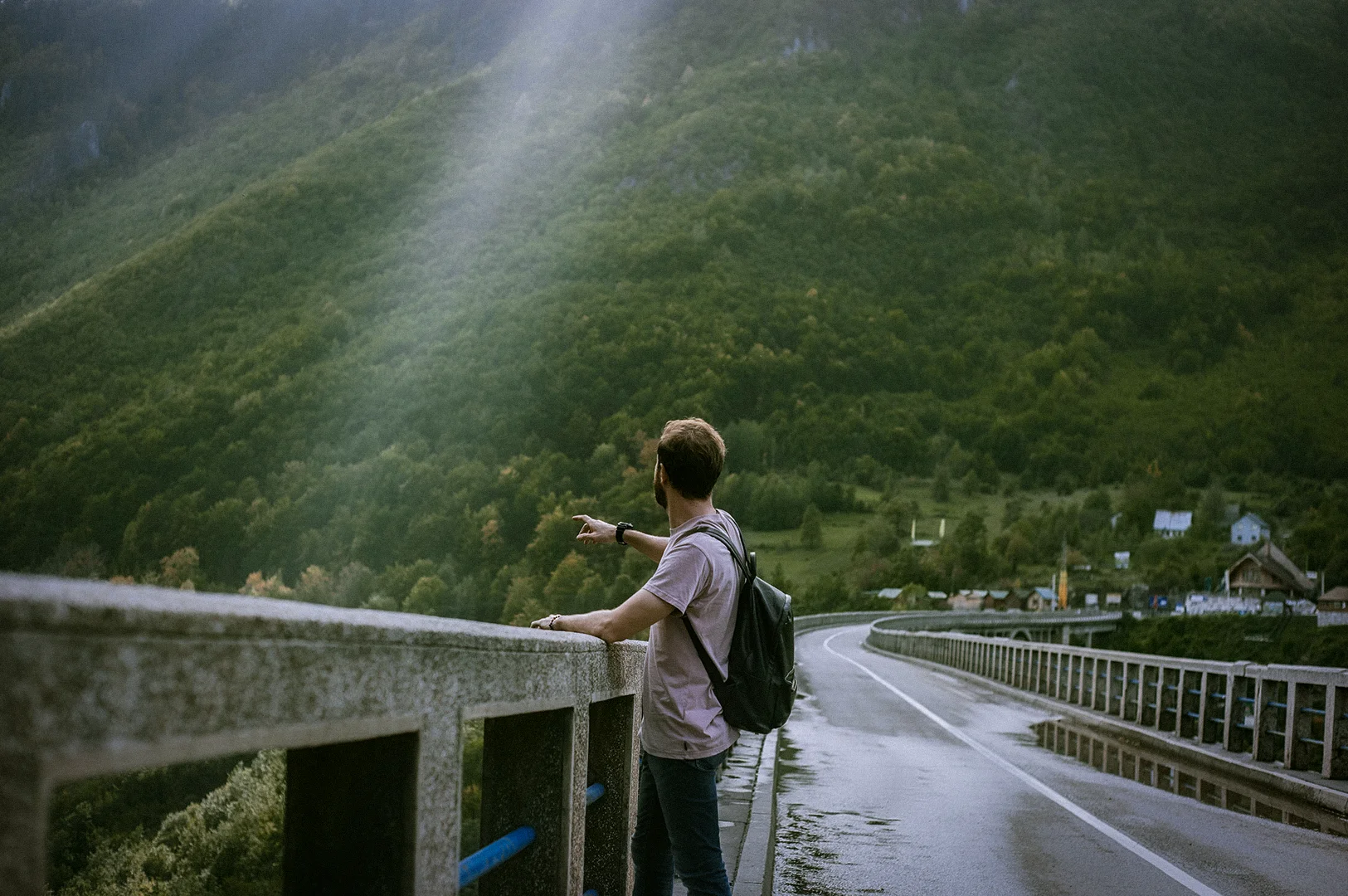 Traveler standing beside a scenic mountain road overlooking lush green hills and forest landscapes during a countryside road trip adventure