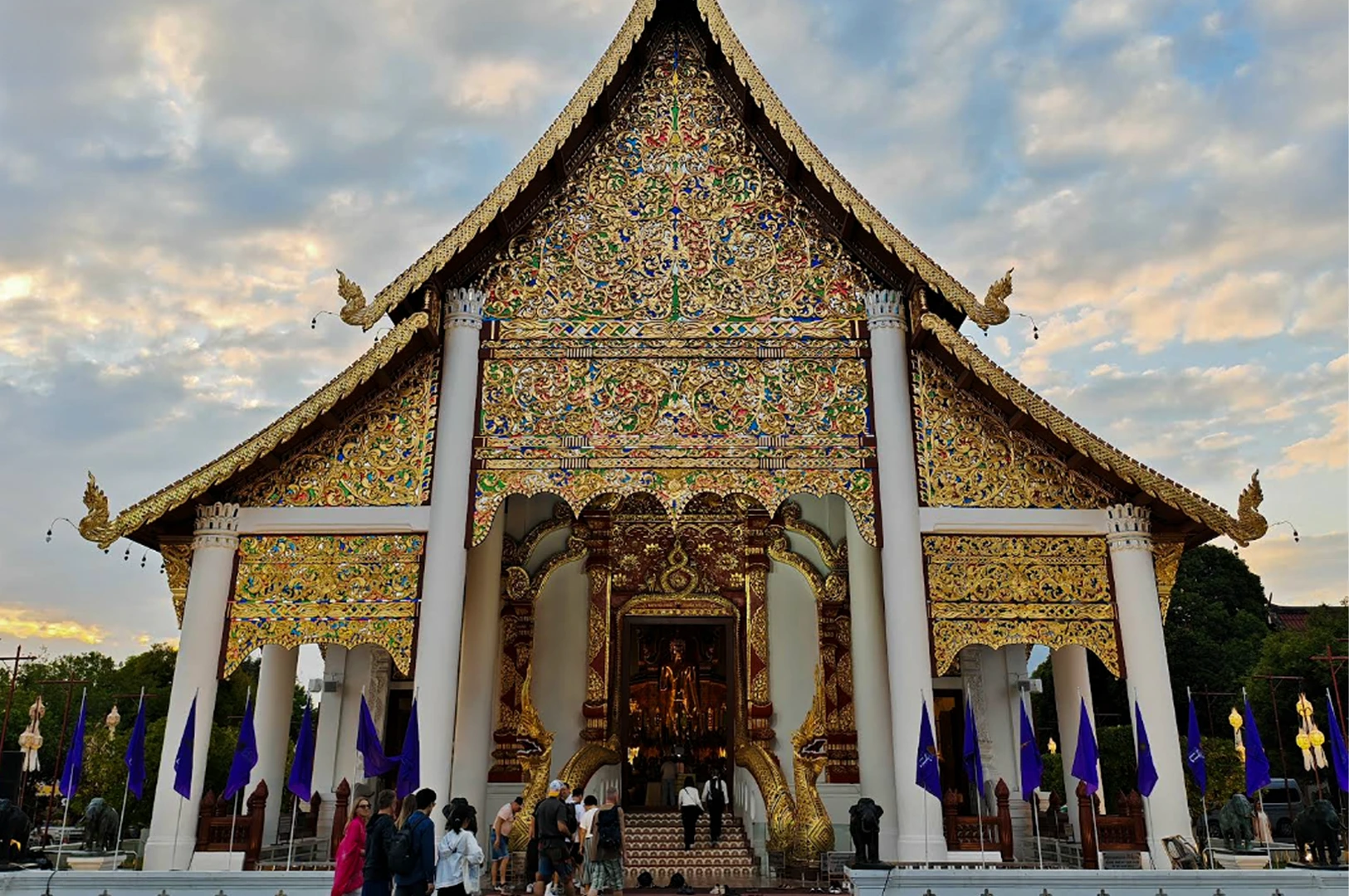 A front-facing view of a magnificent Thai temple (wat) at dusk. The triangular gable is covered in incredibly intricate gold, green, and red filigree. Two large white pillars support the roof, and the entrance is guarded by golden Naga (serpent) statues. A group of tourists stands at the base of the stairs, and several blue flags line the walkway under a soft, cloudy sky.