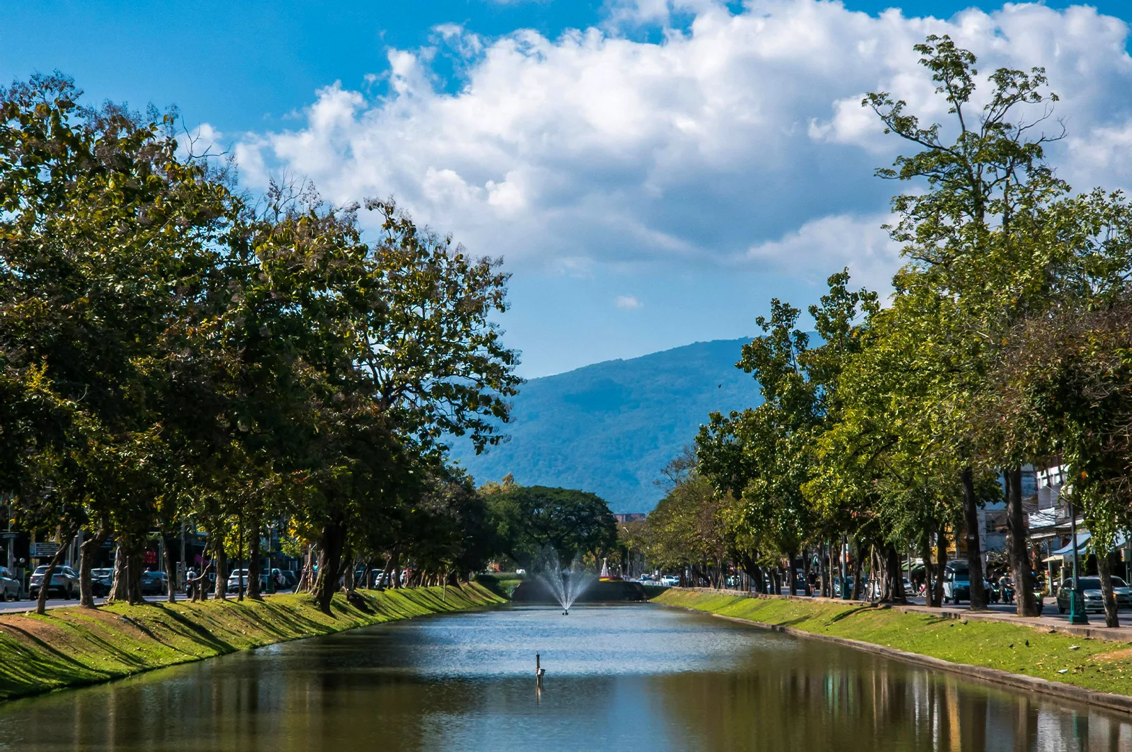 A scenic view looking down a calm, reflective canal in Chiang Mai, flanked by lush green grass banks and rows of leafy trees. In the distance, a large, hazy blue mountain rises against a bright sky filled with soft, white cumulus clouds, and a small water fountain sprays vertically in the center of the canal. The edges of the frame show glimpses of city life, including parked cars and street-side buildings, creating a peaceful contrast between nature and urbanity.