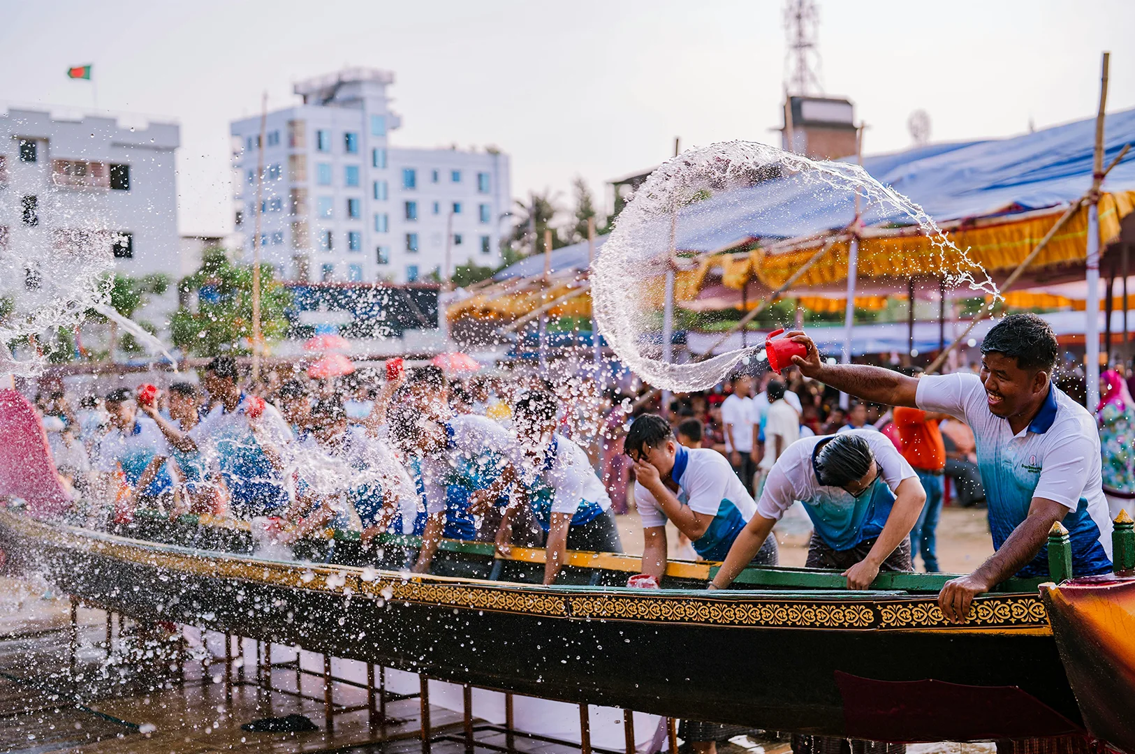 A wide-angle shot of a vibrant community event where a long, dark ceremonial boat with intricate gold patterns sits on a wooden platform. A row of men in matching white and blue gradient shirts stand behind the boat, energetically throwing water from small red buckets. One man in the foreground smiles broadly as he flings a large arc of water into the air. In the background, modern city buildings rise under a pale sky, and a large crowd gathers under yellow and pink awnings.