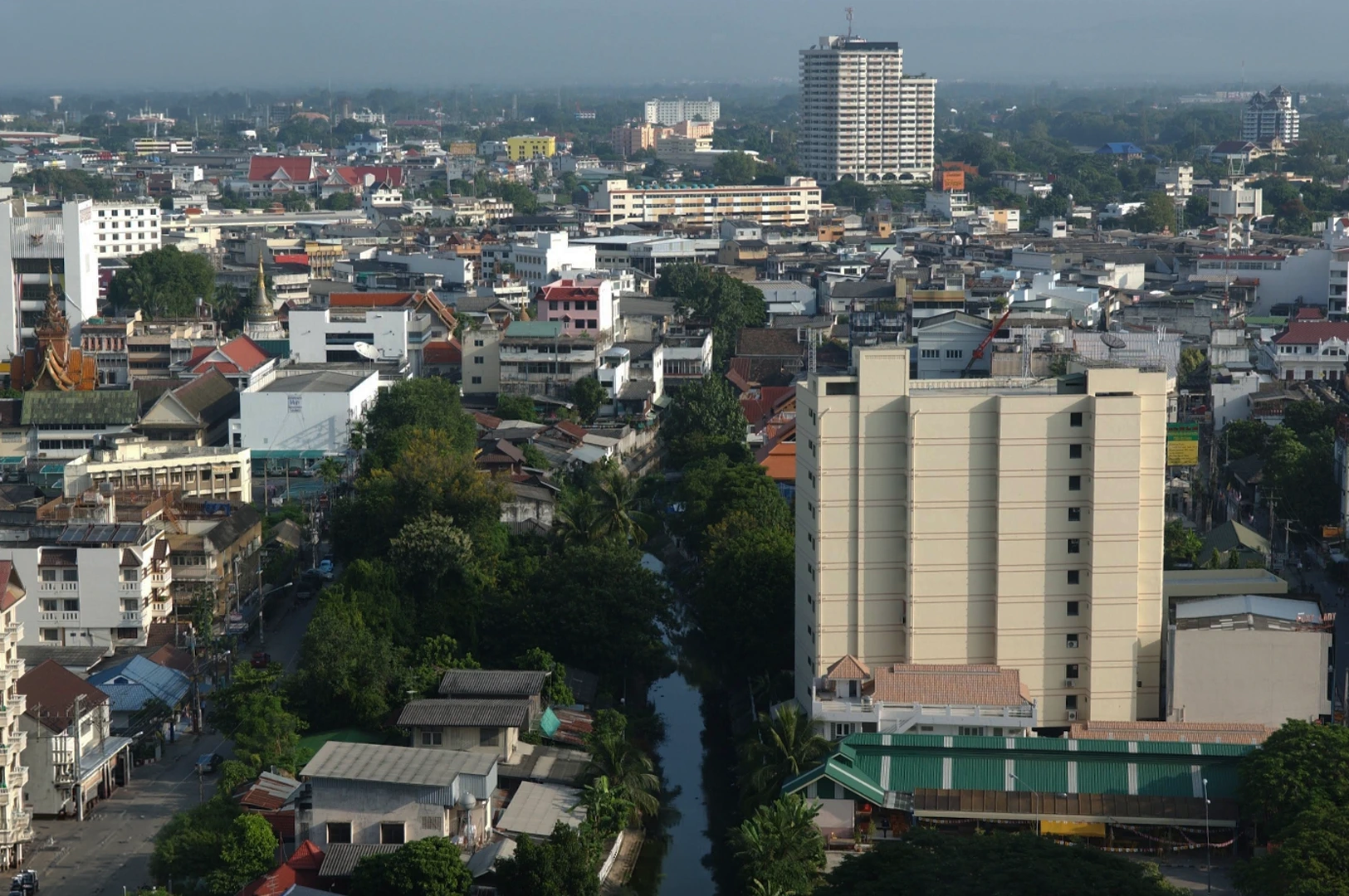 This high-angle cityscape overlooks a densely packed urban area in Chiang Mai, characterized by a mix of low-rise traditional buildings and mid-rise modern structures. A narrow green canal or waterway cuts vertically through the center of the frame, lined with lush tropical trees and vegetation. To the right, a prominent cream-colored rectangular apartment building stands tall, while the background reveals a hazy horizon with a few white skyscrapers rising above a sea of red and grey tiled roofs. The lighting is bright and even, suggesting a clear day.
