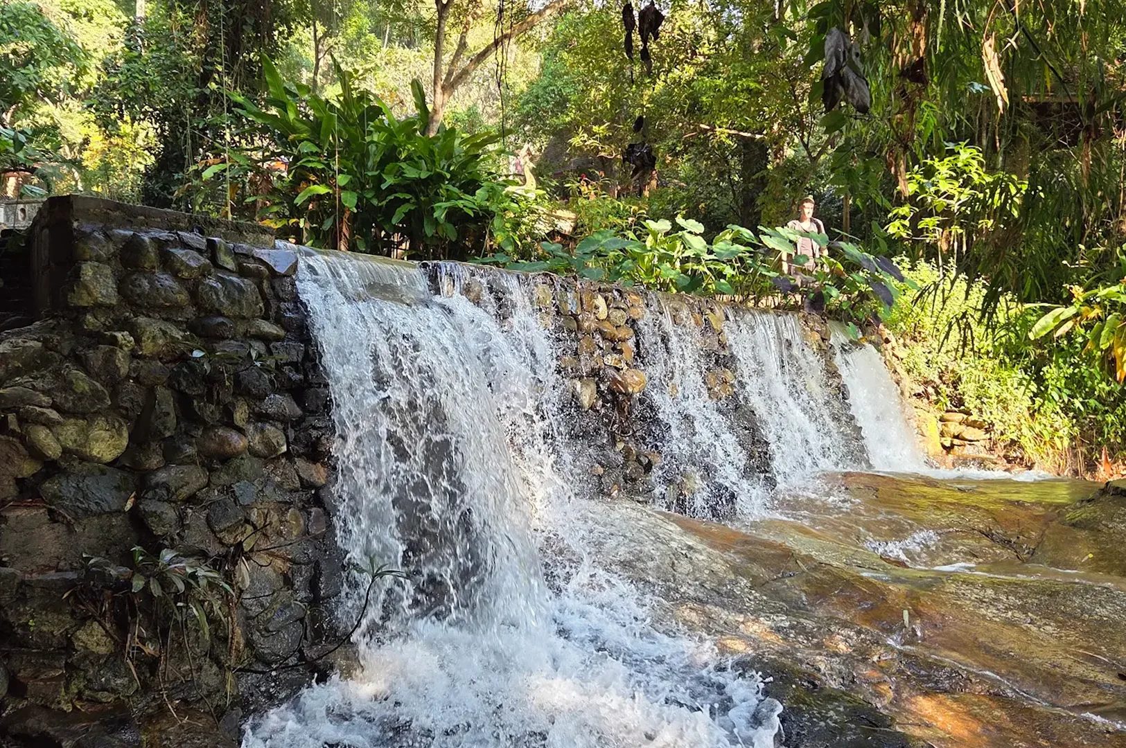 A man-made stone tiered waterfall flowing into a stream surrounded by lush tropical jungle greenery.