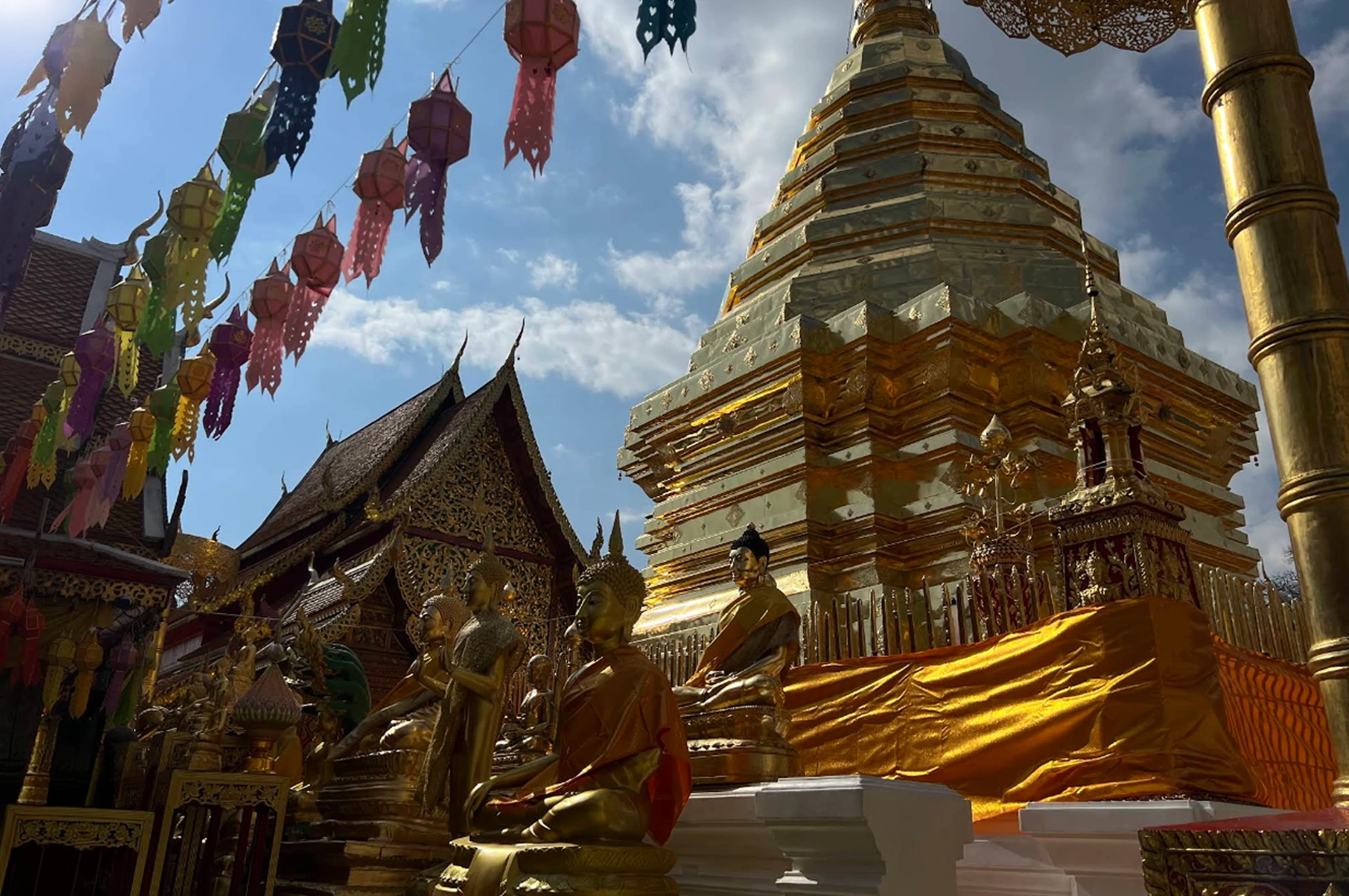 A low-angle shot looking up at a massive, multi-tiered golden stupa reflecting bright sunlight. In the foreground, several serene golden Buddha statues in sitting and standing poses are lined up on a white platform. Colorful paper lanterns in shades of pink, yellow, and blue hang from wires across the top left, set against a bright sky with scattered clouds.