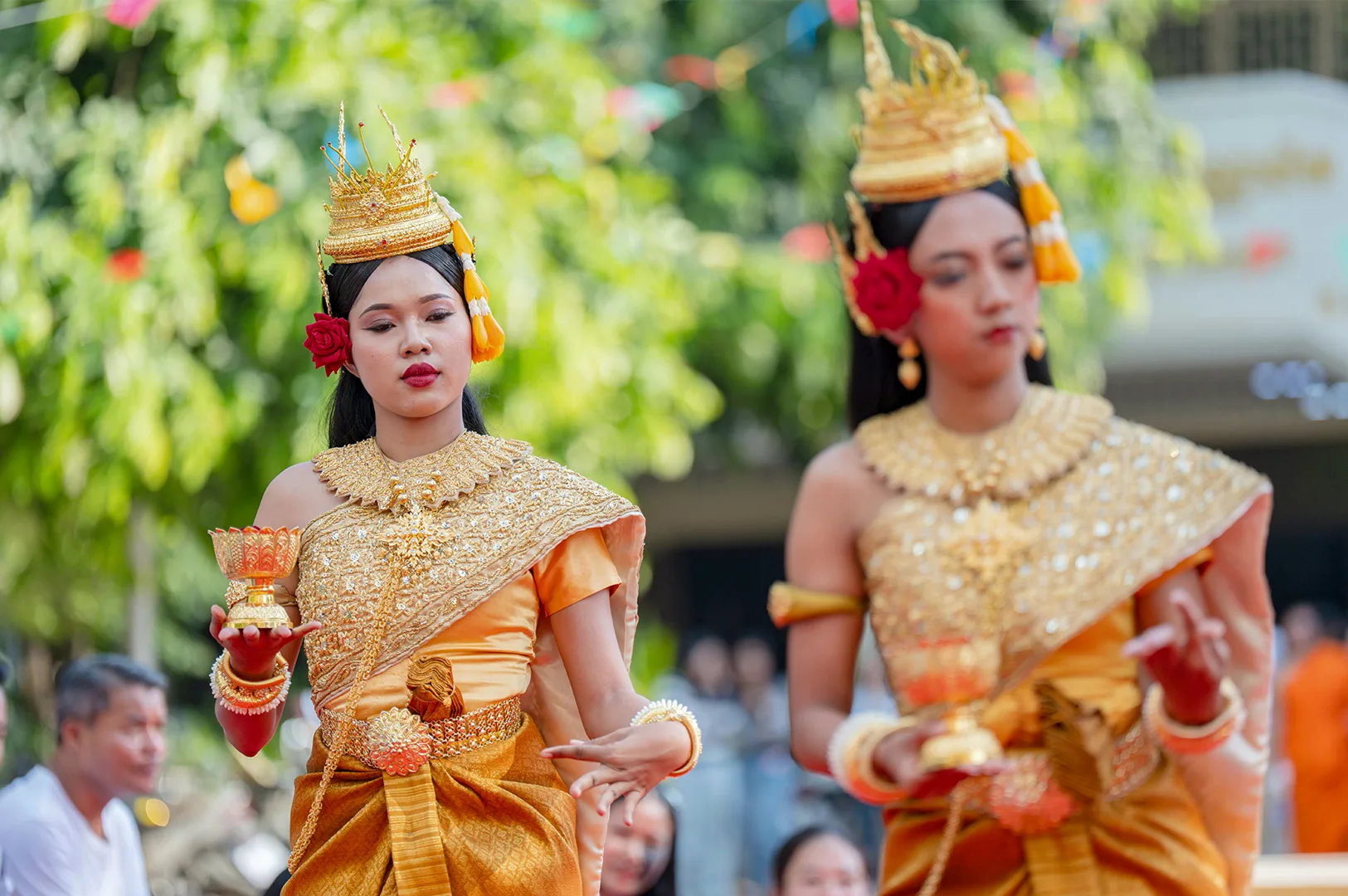 Two women perform a traditional Southeast Asian dance, possibly for Songkran or a similar New Year festival. They are dressed in ornate, shimmering gold costumes with heavy gold necklaces and elaborate peaked crowns decorated with yellow flowers. One dancer in the foreground holds a small gold ceremonial cup, her expression serene and focused. A red flower is tucked behind her ear. The background is a soft blur of green trees and festive decorations.