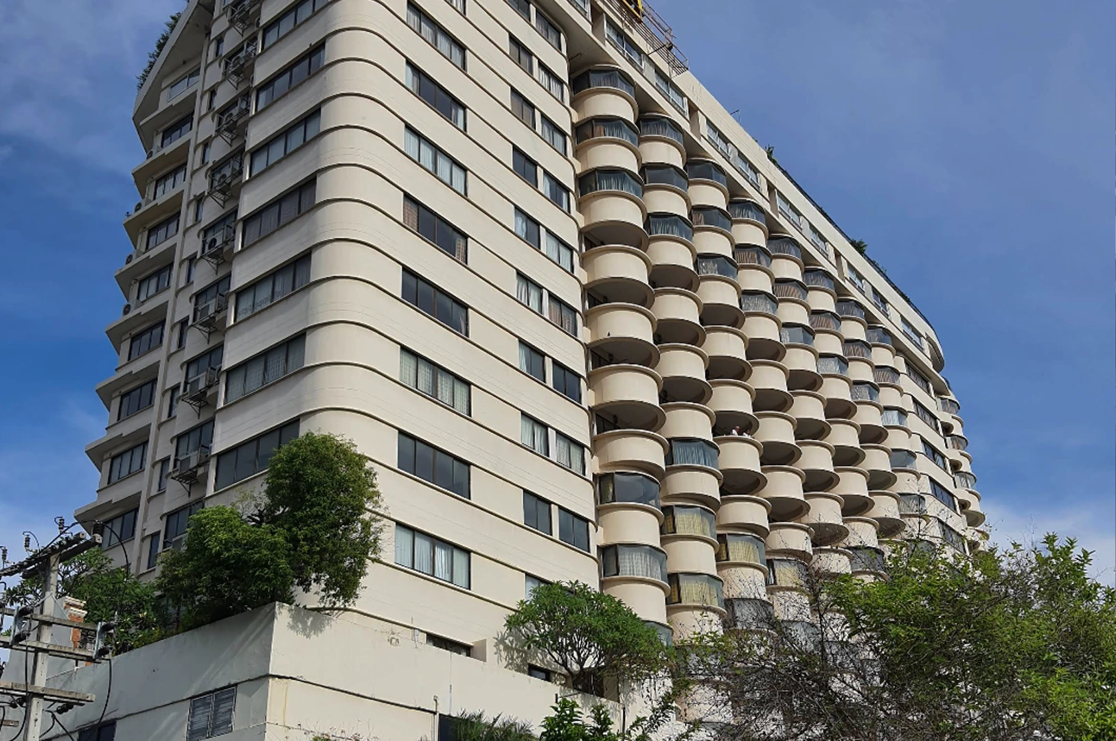 A low-angle shot looks up at a large, distinctive cream-colored apartment complex set against a bright blue sky. The building features a unique architectural design with a vertical stack of rounded, semi-circular balconies on the right side that resemble a honeycomb pattern. The left side of the building has more traditional horizontal windows and flat balconies. Lush green trees and some power lines are visible at the base of the structure, adding a touch of nature to the urban setting.