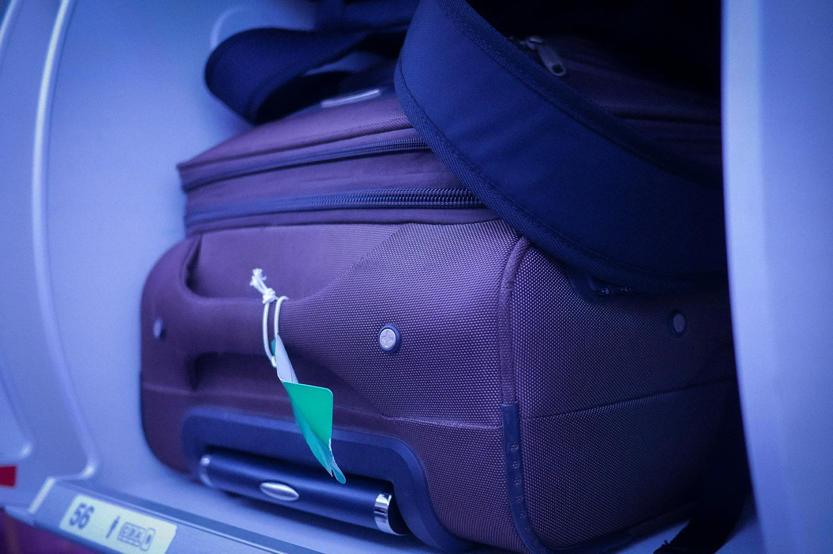 Carry-on suitcase stored inside an airplane overhead compartment with a travel bag and luggage tag during a commercial flight journey