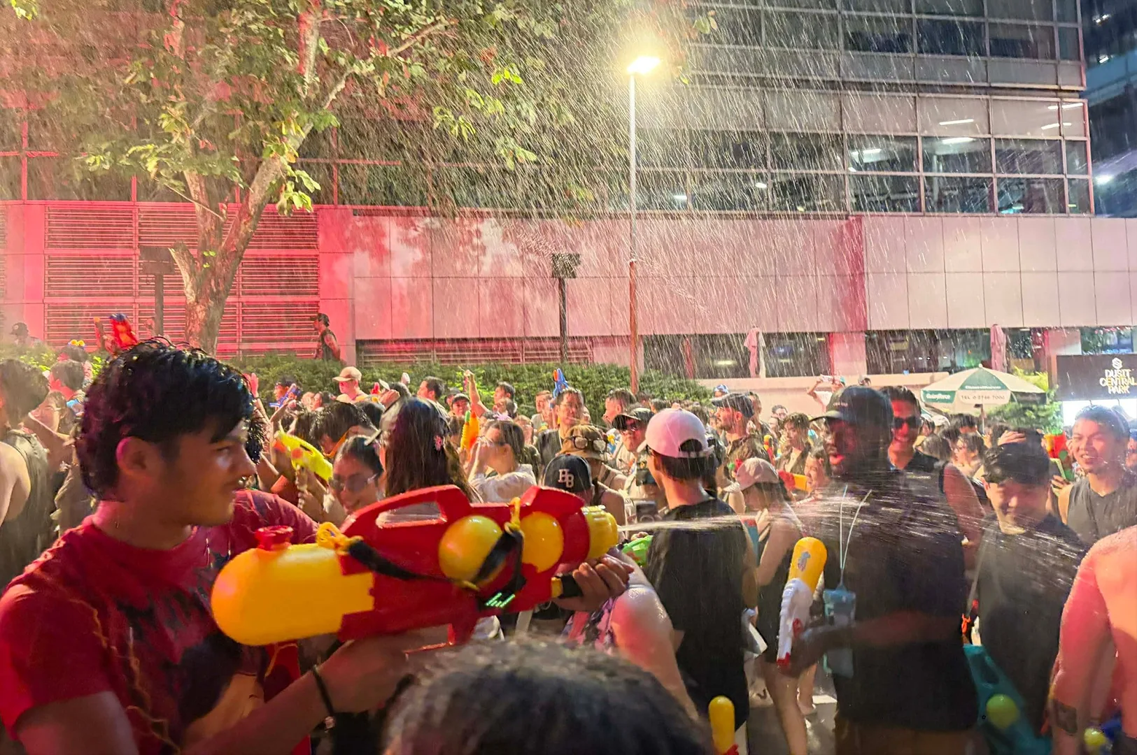 A high-energy nighttime scene of a crowded street water festival. In the foreground, a man in a red shirt aims a large, brightly colored yellow and red water gun. Water sprays are visible everywhere, illuminated by bright overhead streetlights, creating a rain-like effect against the dark sky. The street is packed with people in casual summer clothes, many wearing hats and sunglasses, all participating in the massive, wet celebration in front of a modern pink-toned building.