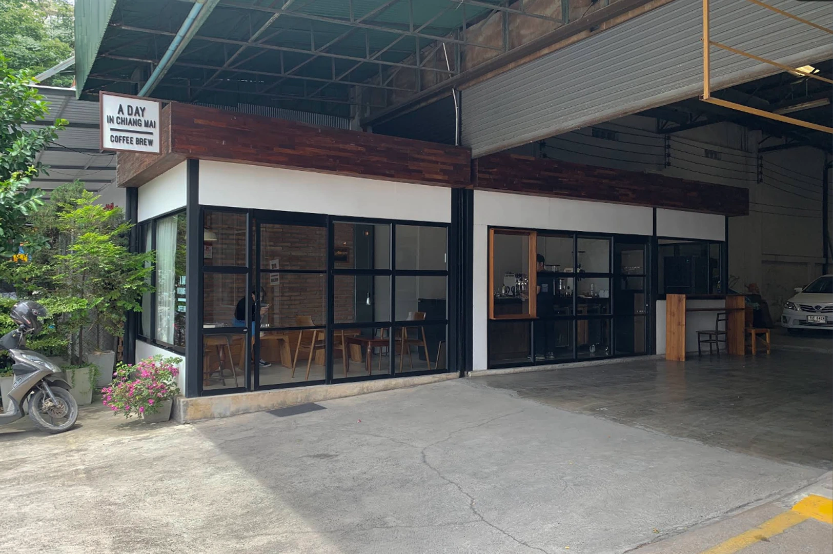 This ground-level shot features the exterior of "A Day in Chiang Mai Coffee Brew," a modern, minimalist cafe located within an industrial-style warehouse space. The cafe is a small, rectangular structure with large glass windows framed in black metal and topped with a dark wood trim. Inside, a patron is visible sitting at a wooden table. The surrounding area consists of a grey concrete floor and a large open garage or warehouse entrance to the right, where a white car is parked in the shadows.