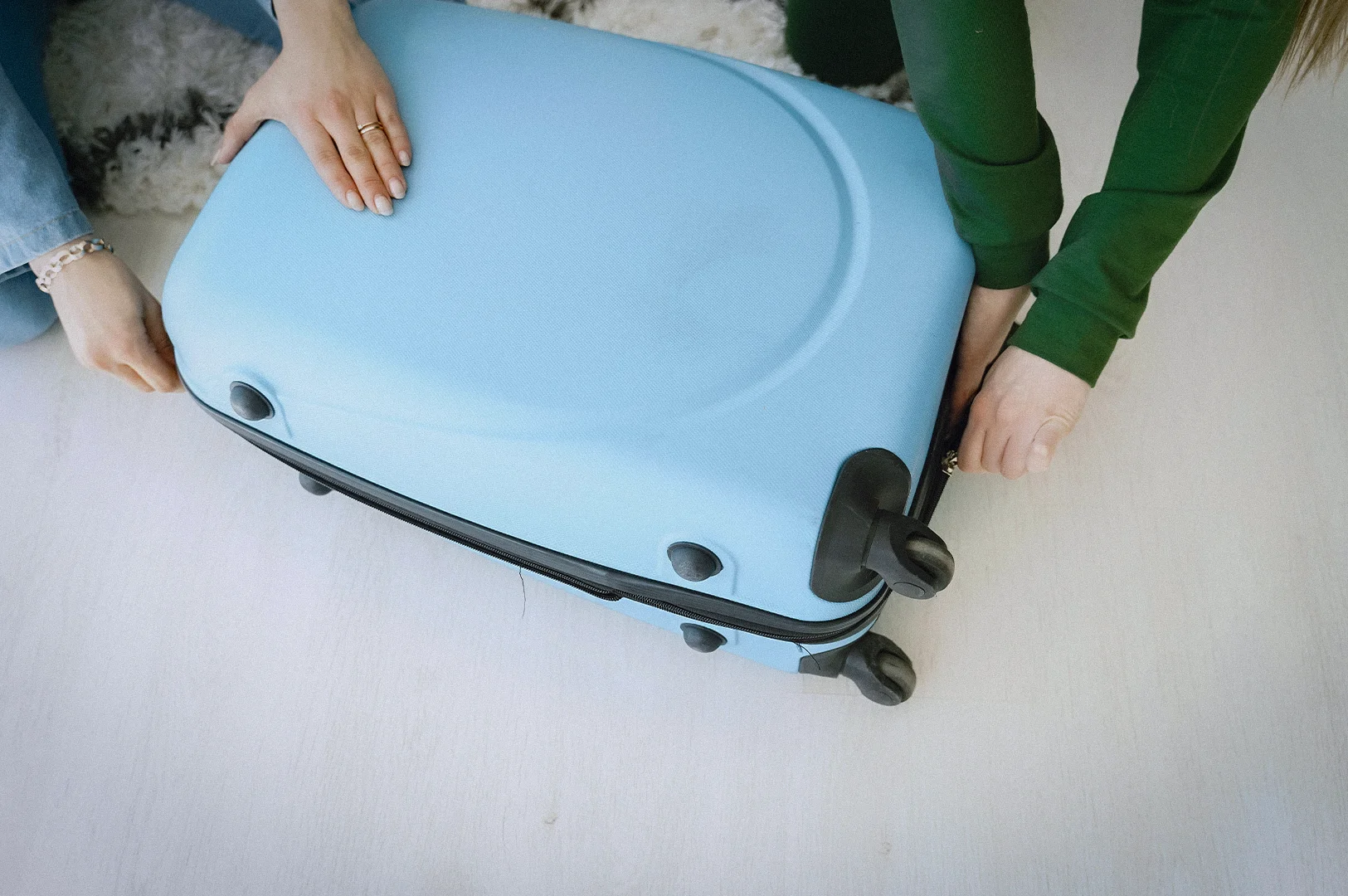 Two people closing an overpacked blue hard shell suitcase while preparing luggage for travel at home on a light wooden floor