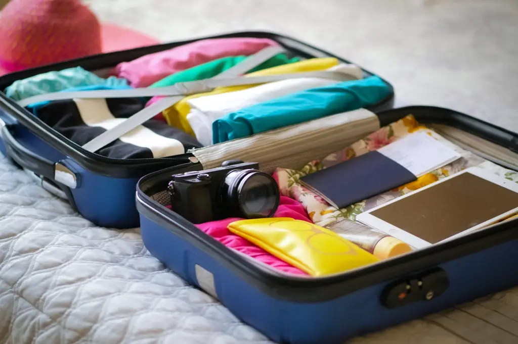 Open travel suitcase packed with colorful clothes, camera, passport, tablet, and vacation essentials on a bed, ready for a holiday trip or travel adventure