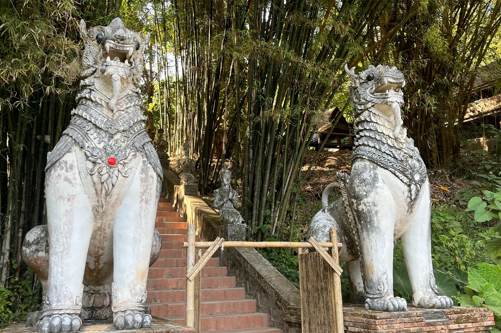 Two large white Singha Mythical Lion guardian statues standing at the base of temple stairs in a bamboo forest.