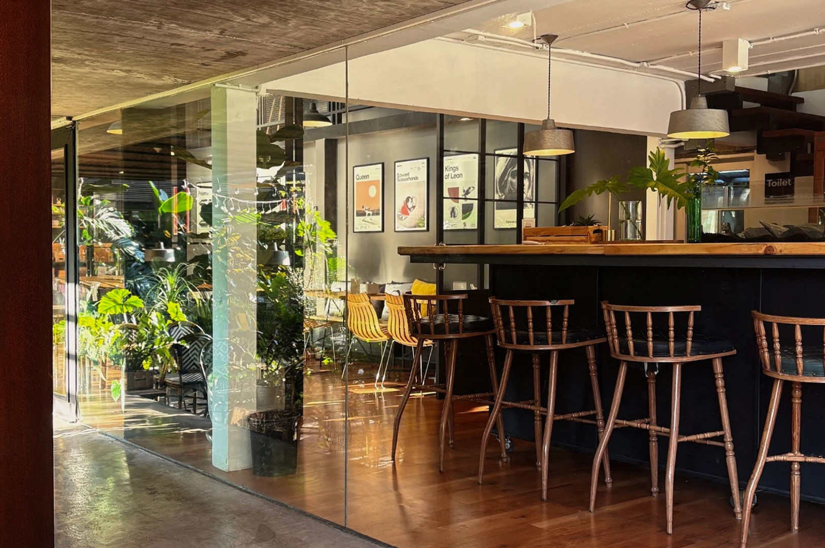An interior view of a stylish, modern cafe with warm wood floors and a black service counter. High-backed wooden bar stools are tucked under the counter. A large glass wall separates the indoor area from an outdoor patio filled with lush green tropical plants. On the gray interior wall, four minimalist posters are framed, and industrial-style pendant lights hang from the ceiling.