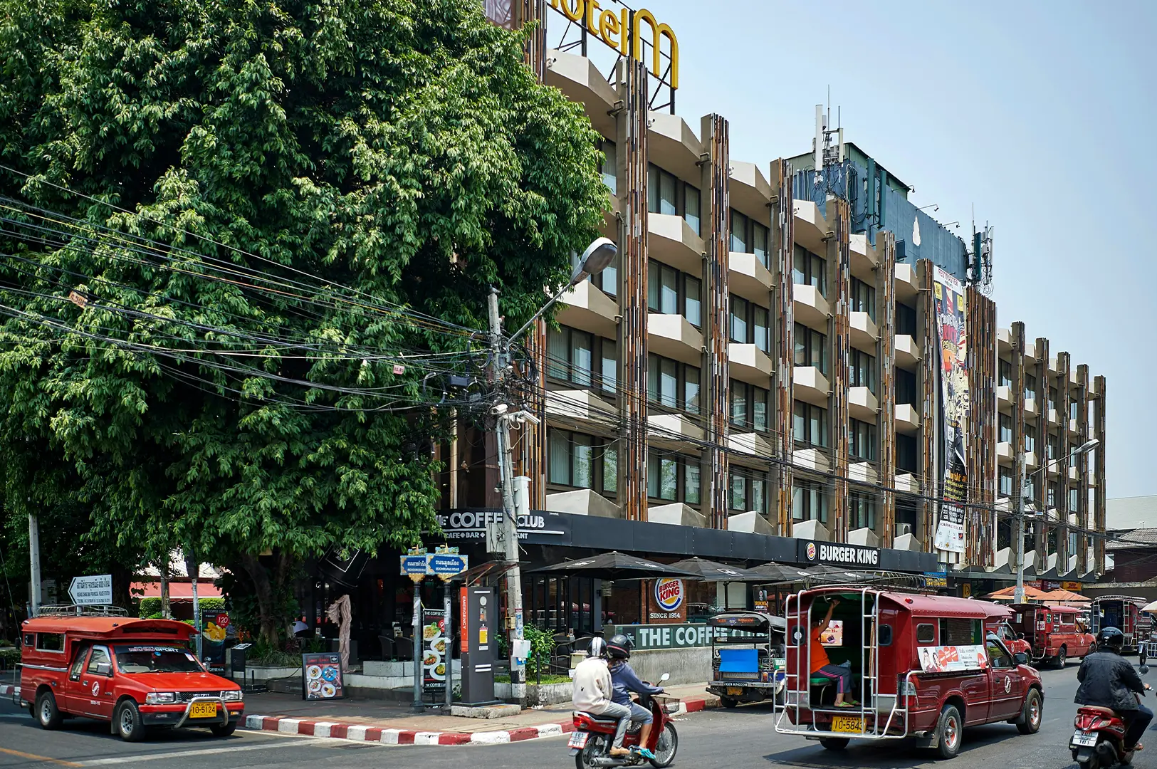 A busy street corner in Chiang Mai featuring a multi-story hotel building with a unique facade of vertical wooden slats and white balconies. At the street level, familiar global franchises like Burger King and The Coffee Club are visible beneath a dense canopy of a large green tree and a chaotic web of overhead power lines. Red "songthaew" passenger trucks and several motorbikes navigate the intersection, capturing the warm, sunlit atmosphere of a typical afternoon in the city.
