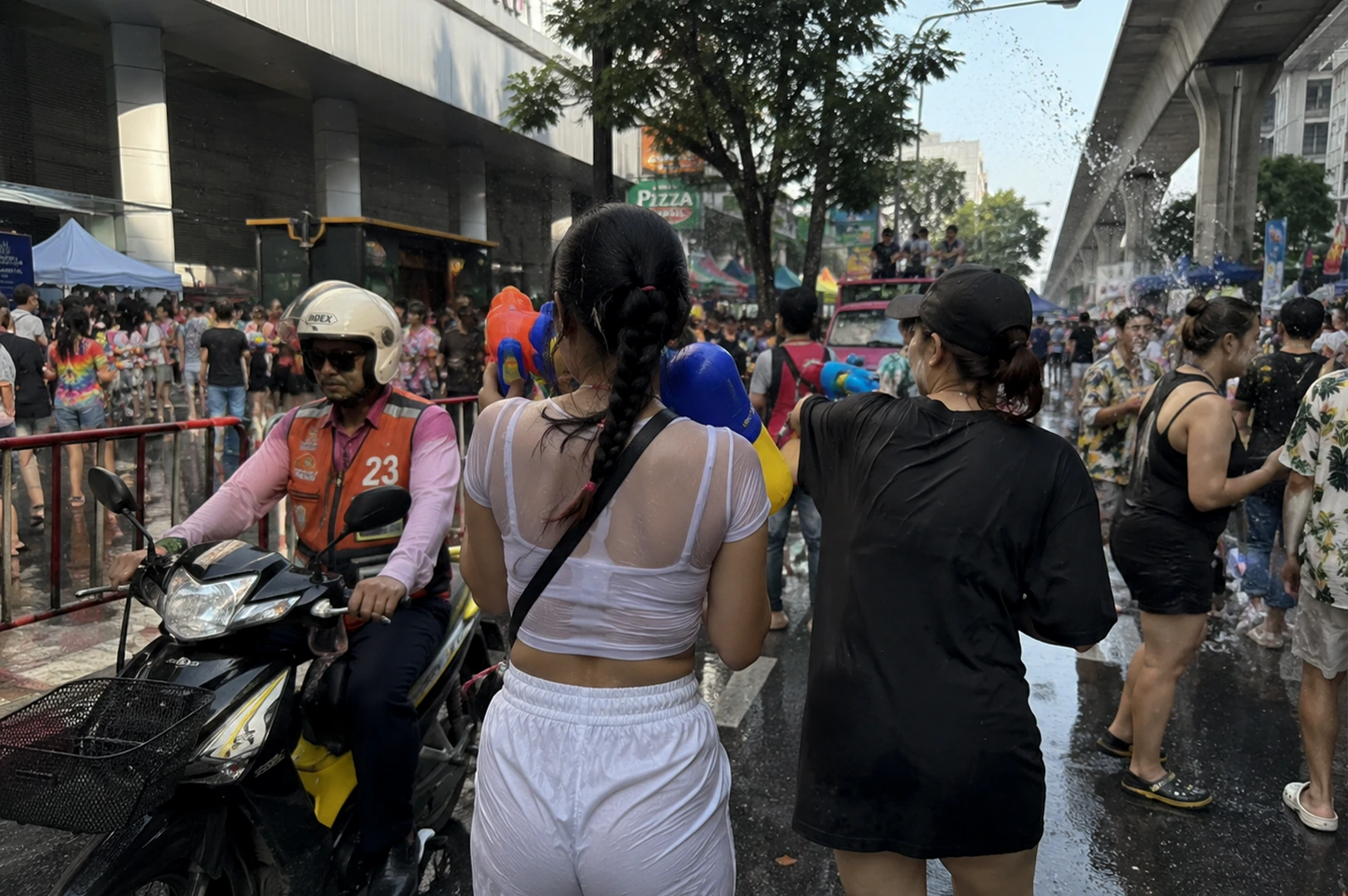 A street-level view of a water festival in a busy city. Two women stand in the center, their backs to the camera; one wears a white mesh top and white shorts, while the other is in a black t-shirt. Both carry large, colorful water blasters. To the left, a man on a yellow motorcycle wearing a helmet and a bright orange vest (number 23) navigates through the wet street. The pavement is soaked, and the background shows city infrastructure, including a pedestrian overpass and urban storefronts.