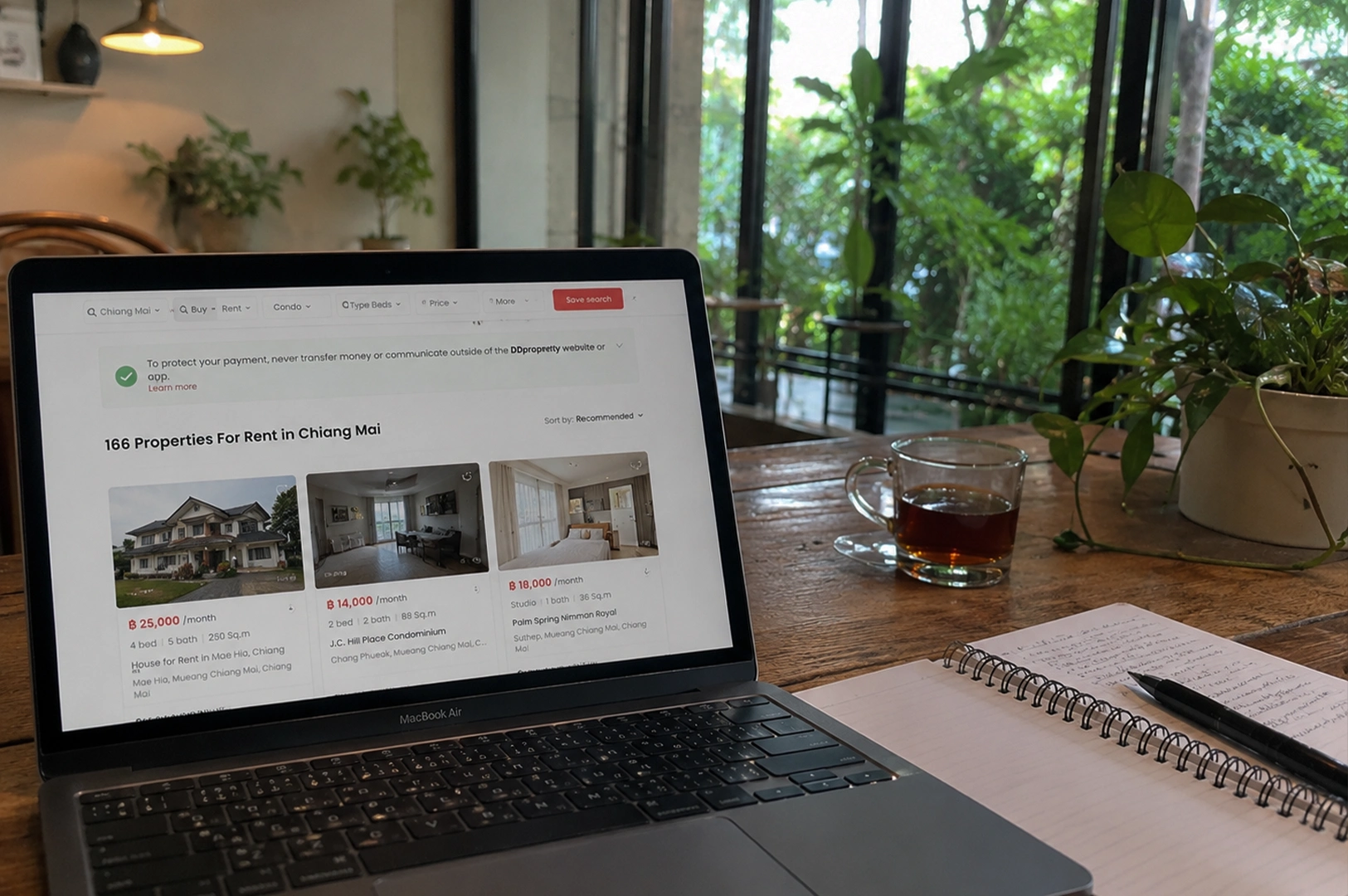 A cozy, indoor lifestyle shot shows a silver laptop open on a rustic wooden table, displaying a real estate website titled "166 Properties For Rent in Chiang Mai." Next to the laptop sits a glass mug of dark tea and a spiral-bound notebook with a black pen resting on it. The background is softly blurred, revealing large glass windows that look out onto a vibrant green garden, creating a peaceful "digital nomad" workspace atmosphere filled with natural light and indoor plants.
