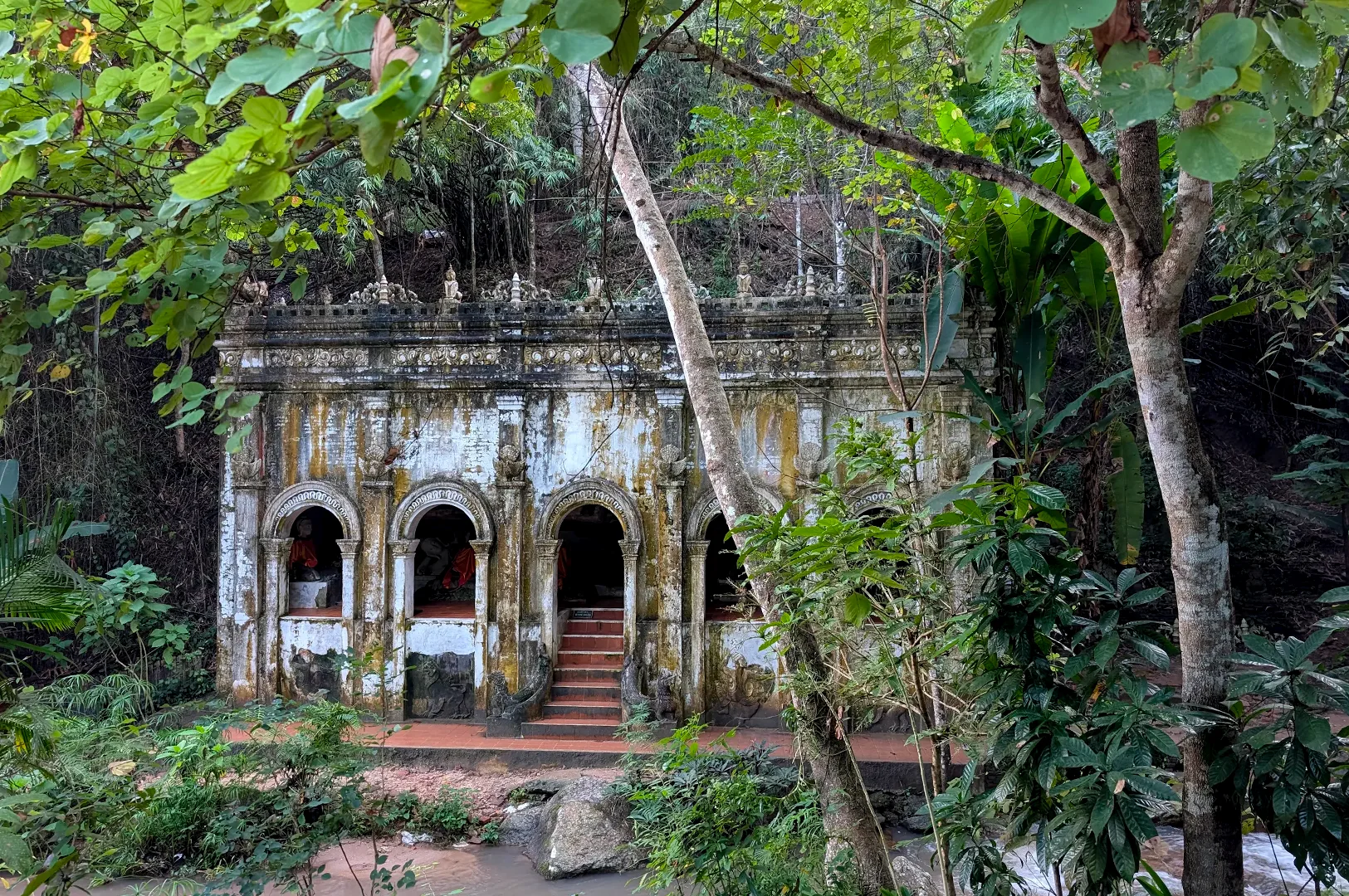 An weathered ancient stone temple structure with arched openings hidden within a dense tropical forest.