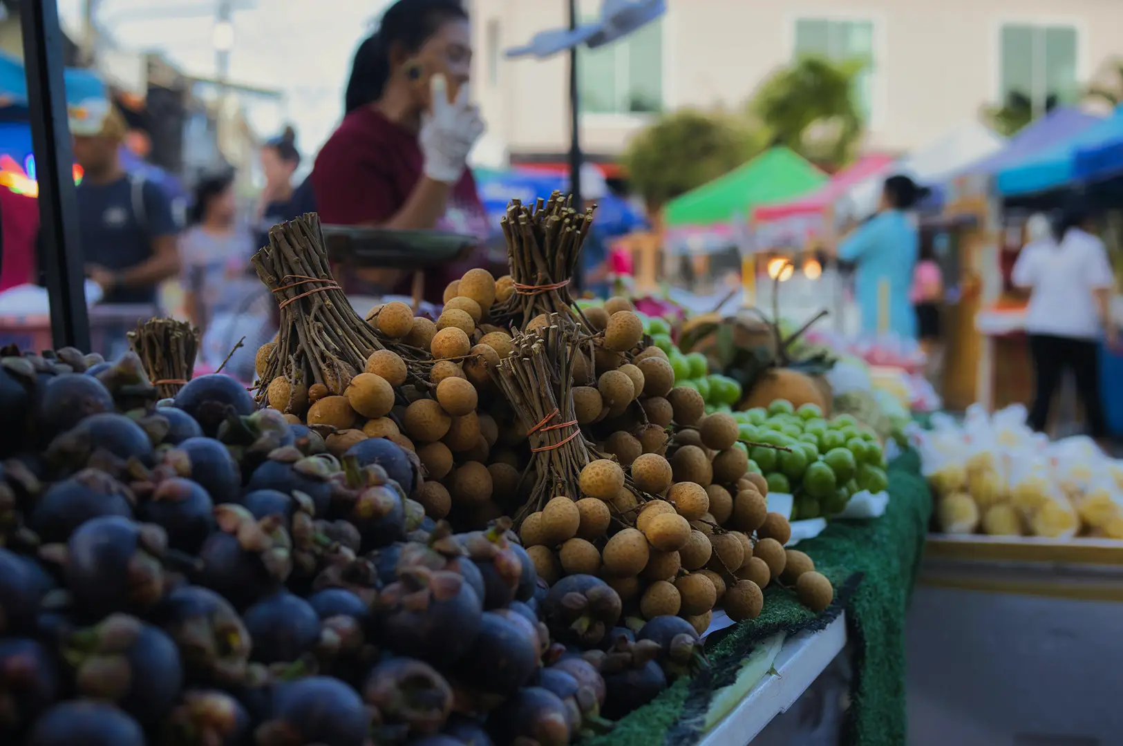A close-up, shallow-depth-of-field shot of a fruit stall at an outdoor market, focusing on bunches of brown, round longan fruit tied together with rubber bands. To the left are dark purple, thick-skinned mangosteens, and in the background, out-of-focus green grapes and other produce are visible. Blurred figures of vendors and shoppers move behind the display under the soft light of market tents, emphasizing the freshness and variety of local tropical fruits.