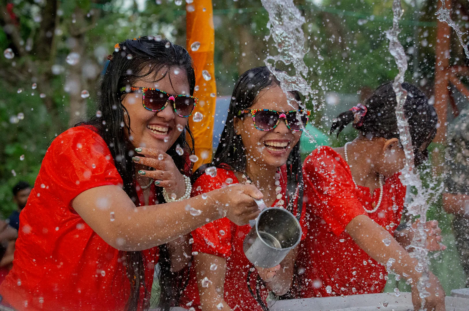 Three girls in matching bright red shirts are captured in a moment of pure laughter and fun during a water celebration. The two girls in the foreground wear colorful, patterned sunglasses and are drenched. One girl holds a small metal cup, splashing water toward the camera. Large, clear water droplets are scattered across the entire frame, catching the light. Their expressions are exuberant, and one girl has a pearl necklace and decorative henna on her hand.