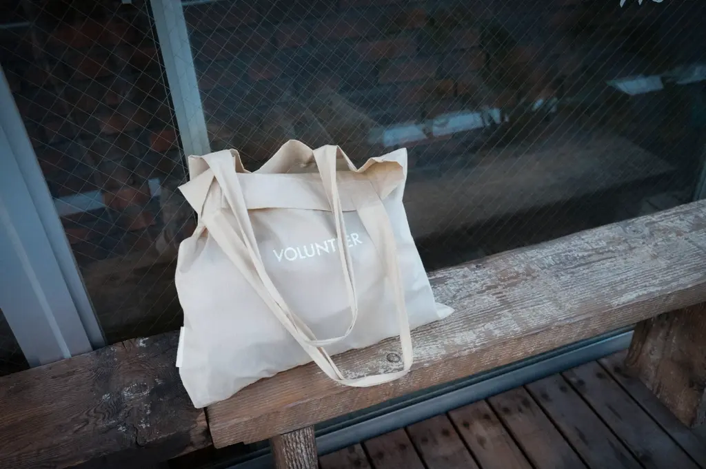 Minimalist canvas tote bag with “Volunteer” text placed on a rustic wooden bench beside a glass window in an urban outdoor setting