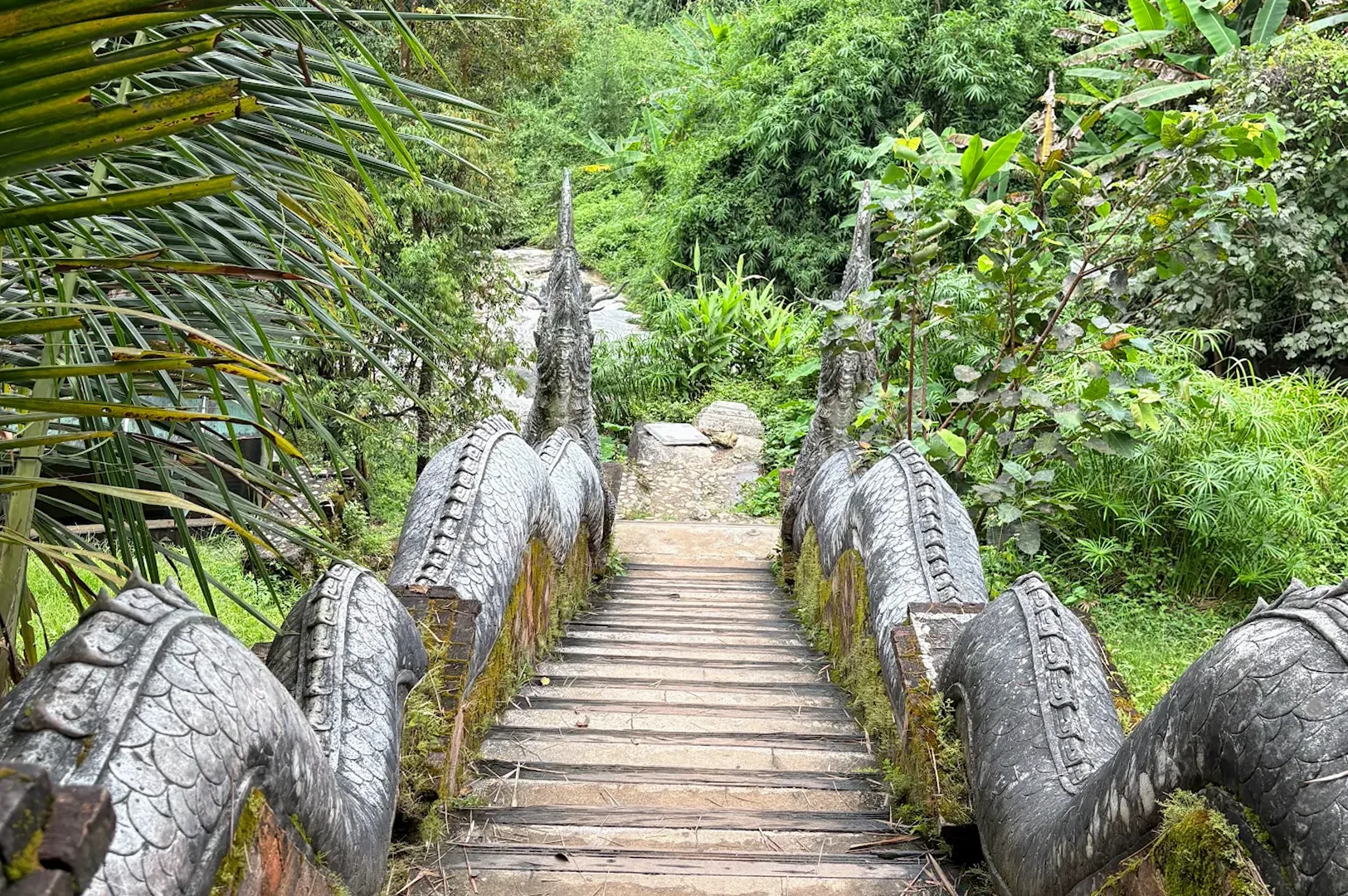 A stone staircase guarded by Naga serpent banisters leading down towards a waterfall in a lush jungle setting.