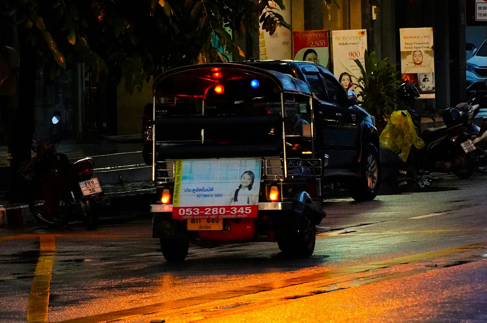 A night scene on a wet city street, showing the back of a traditional three-wheeled tuk-tuk driving away. The asphalt is slick with rain, reflecting the warm orange glow of streetlights and the tuk-tuk’s red and blue tail lights. In the background, posters for beauty services and a parked black SUV are visible, while the overall mood is dark and atmospheric, highlighting the reflective surfaces and colorful lights of Chiang Mai after a rainfall.