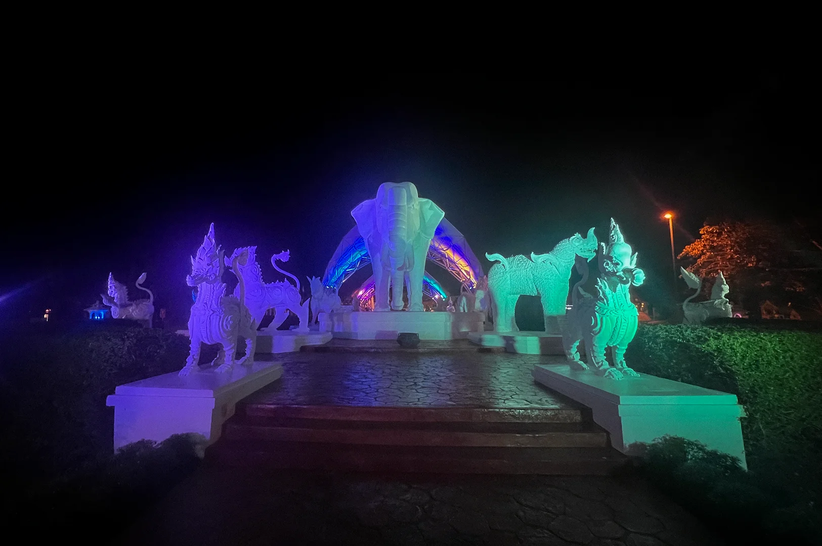 Illuminated elephant statue and traditional mythical creature sculptures at the entrance of Chiang Mai Night Safari in Thailand during nighttime with colorful lights and landscaped pathways