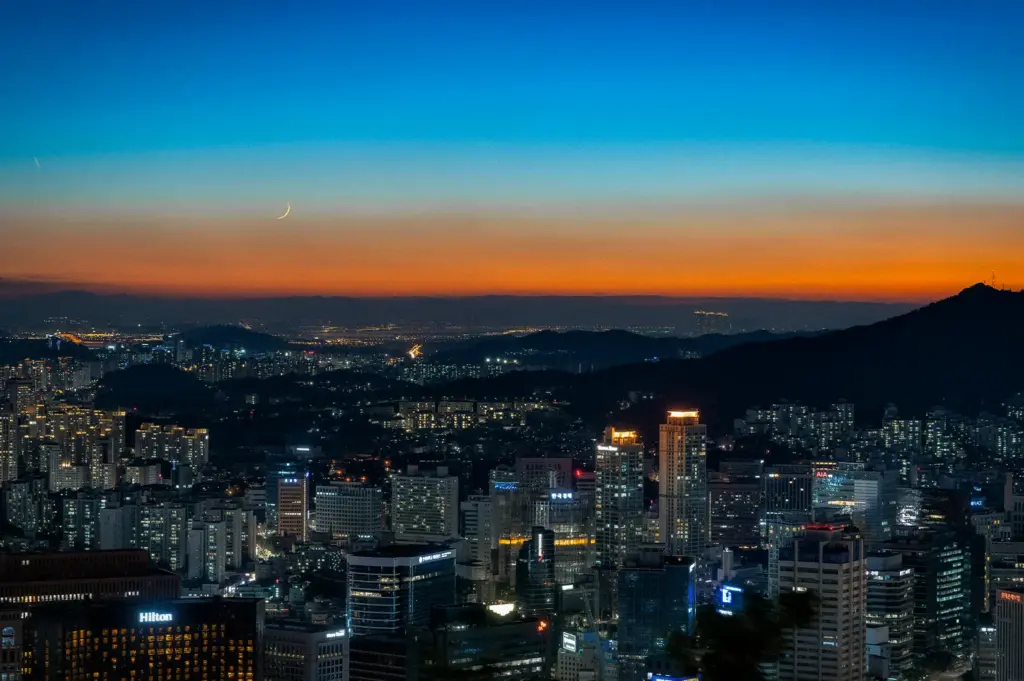 Panoramic city skyline at twilight with illuminated skyscrapers, distant hills, and a crescent moon above the horizon, capturing a vibrant urban landscape transitioning from sunset to night.