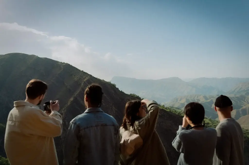 Group of travelers admiring a panoramic mountain landscape from a scenic viewpoint, capturing photos and enjoying an outdoor adventure in a rugged natural setting.