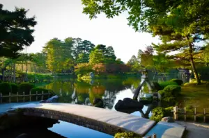 Traditional Japanese garden featuring a stone bridge, tranquil pond, landscaped trees, and reflective water in a scenic outdoor setting.