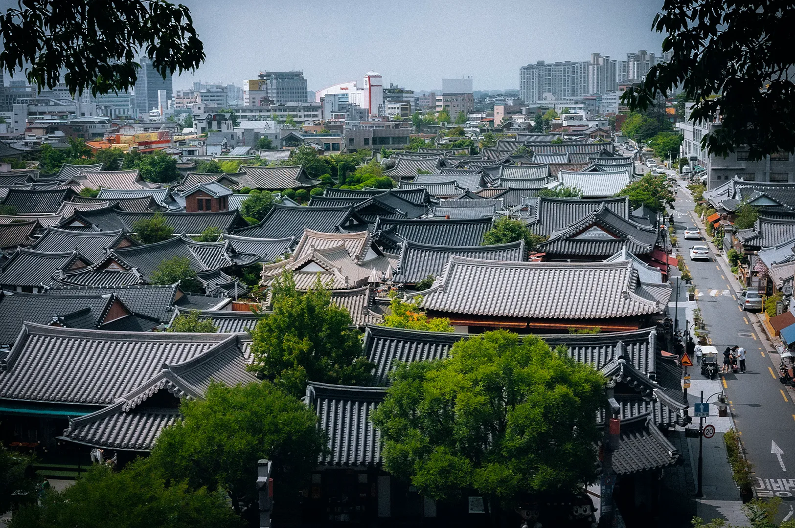 Panoramic view of Jeonju Hanok Village in South Korea featuring traditional Korean hanok rooftops, tree-lined streets and the modern city skyline in the background.