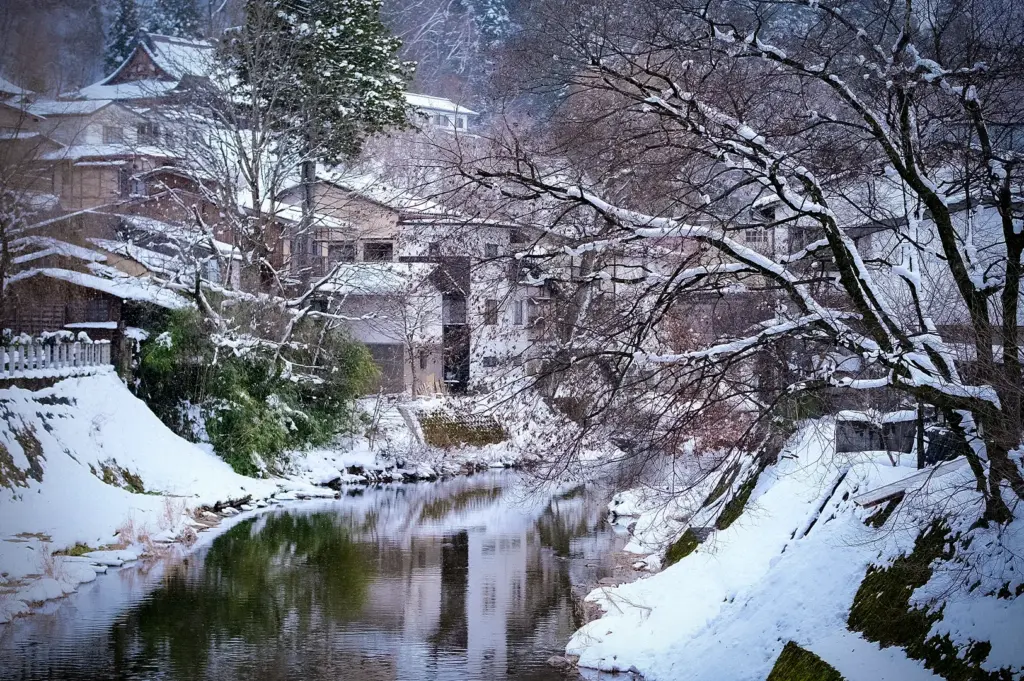 Snow-covered river flowing through Takayama, Japan, with traditional houses, bare winter trees, and peaceful reflections creating a scenic alpine winter landscape.