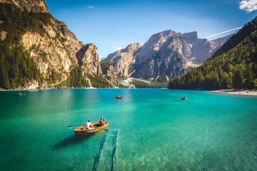 Crystal-clear turquoise alpine lake surrounded by dramatic mountains and pine forests with people rowing wooden boats on a sunny day