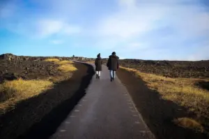 Two people walking along a paved trail through a rugged volcanic landscape with rocky terrain, dry grass, and expansive blue skies during a scenic outdoor hike.