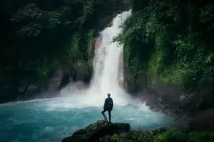 Traveler standing on a rock overlooking a powerful waterfall surrounded by lush tropical rainforest, misty blue waters, and dense green jungle scenery.
