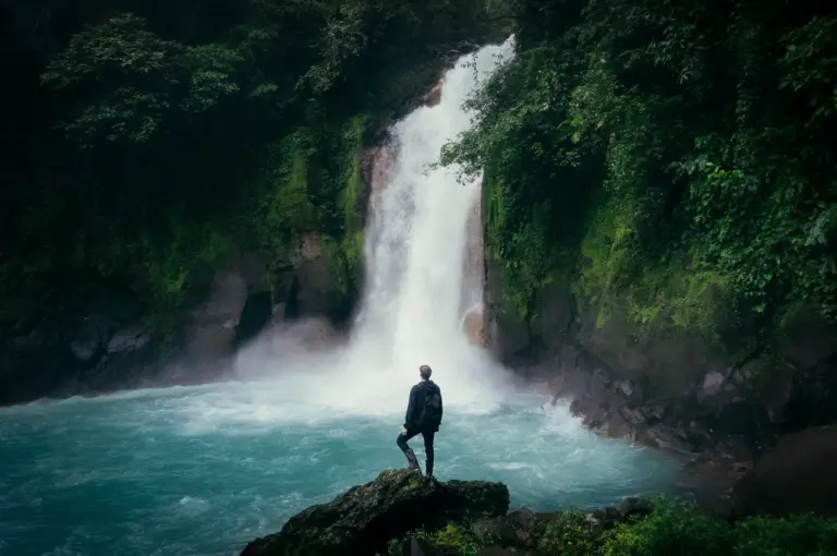 Traveler standing on a rock overlooking a powerful waterfall surrounded by lush tropical rainforest, misty blue waters, and dense green jungle scenery.