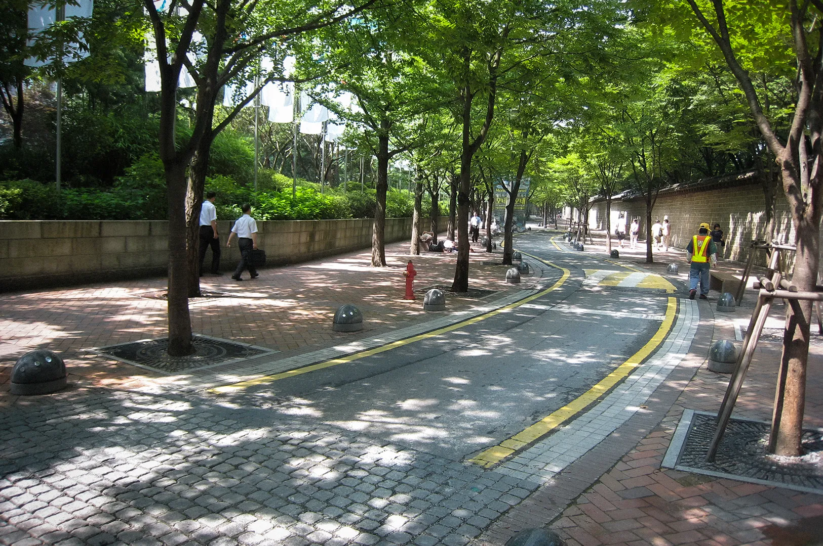 Shaded pedestrian street in Seoul lined with mature green trees, cobblestone walkways, and a gently curving road. People stroll beneath the leafy canopy on a bright summer day, showcasing Seoul’s walkable urban landscape and peaceful city atmosphere.