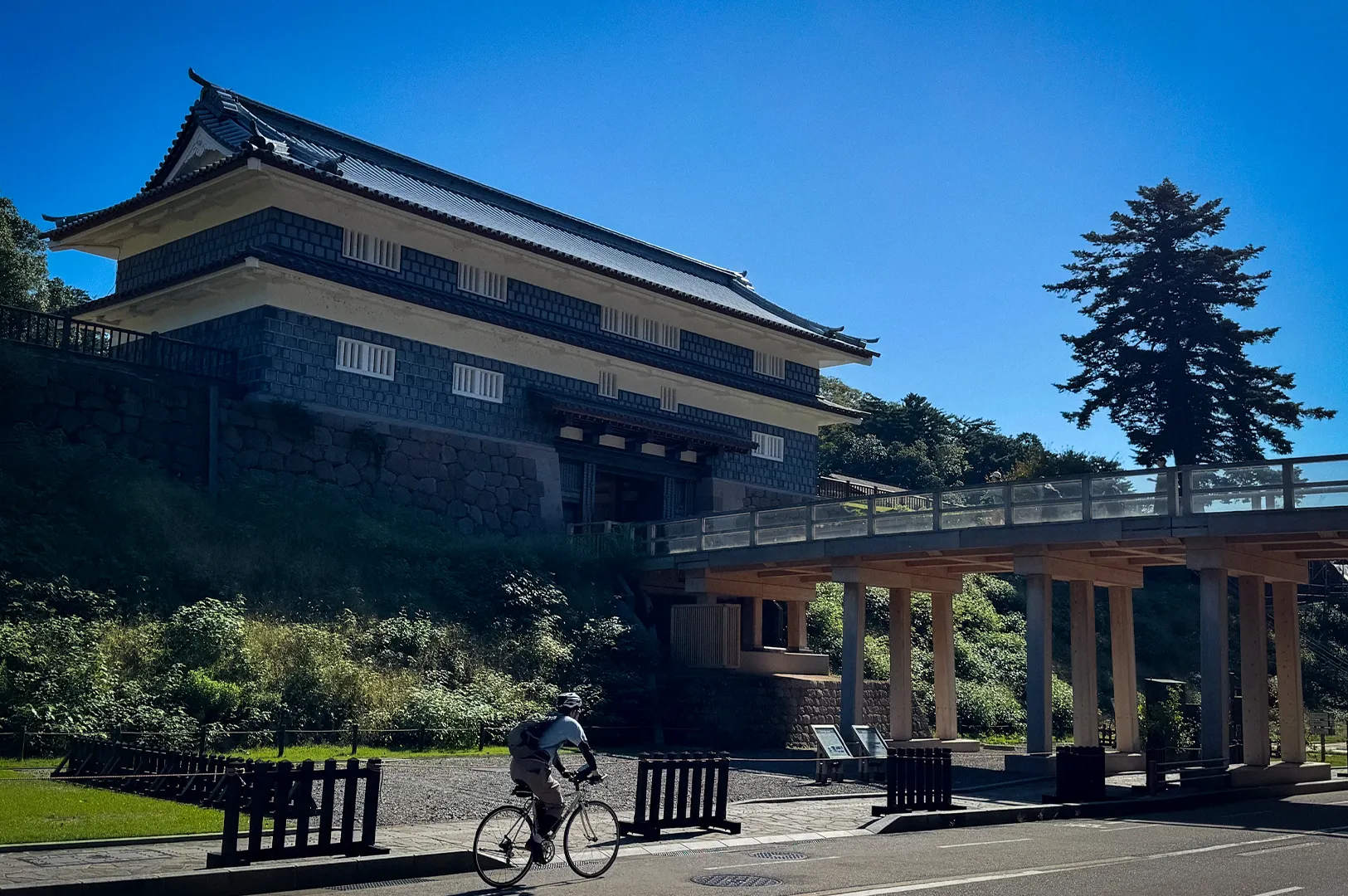 Historic gate structure of Kanazawa Castle in Kanazawa, Japan, featuring traditional white-and-black castle architecture, stone walls, and a cyclist passing by on a sunny day beneath a clear blue sky.