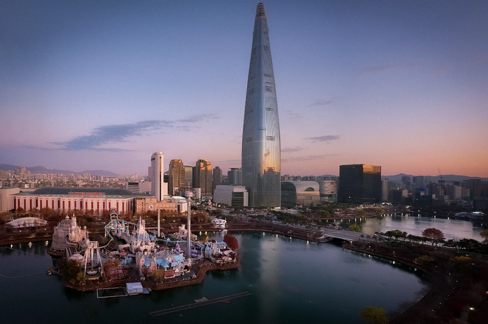 Lotte World Tower rising above the Seoul skyline at dusk, overlooking Seokchon Lake and Lotte World theme park. The iconic skyscraper dominates the cityscape with modern high-rise buildings, calm waterfront views, and soft evening light in South Korea.