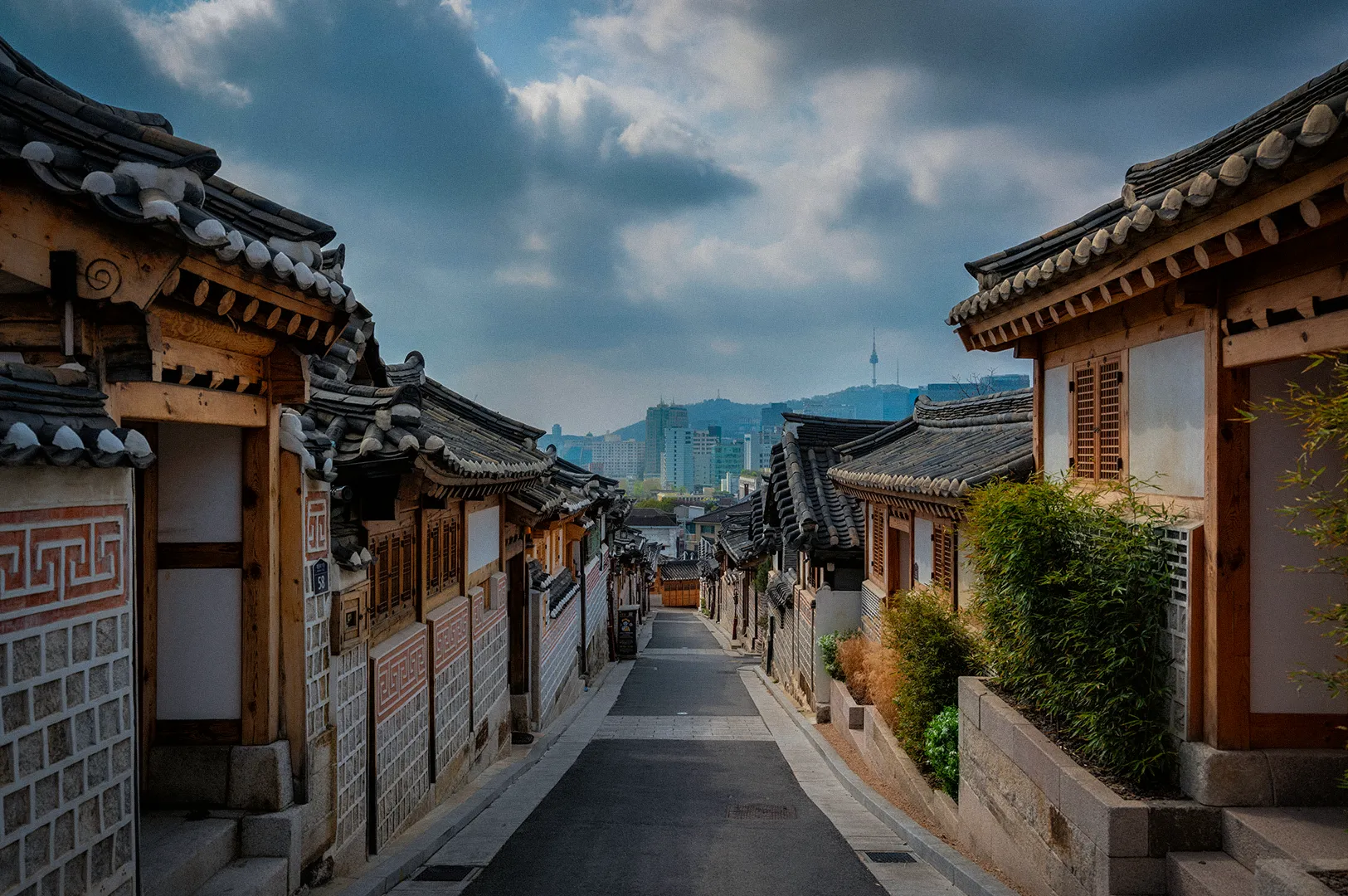 Traditional Korean hanok houses lining a quiet street in Bukchon Hanok Village, Seoul. The historic neighborhood features tiled rooftops, wooden architecture, and views of the modern Seoul skyline with N Seoul Tower visible in the distance beneath dramatic clouds.