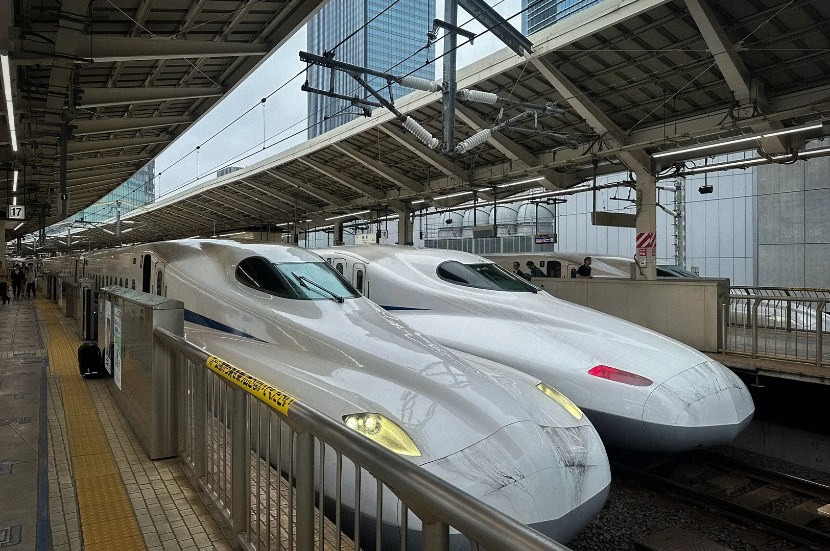 Two Shinkansen bullet trains are stationed side by side at Tokyo Station in Japan, with passengers waiting on the platform beneath the covered railway terminal.