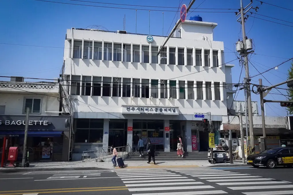 Front view of Jeonju Express Bus Terminal in Jeonju, South Korea, featuring the terminal building, pedestrian crosswalk, nearby shops, and travelers entering and exiting the transport hub.